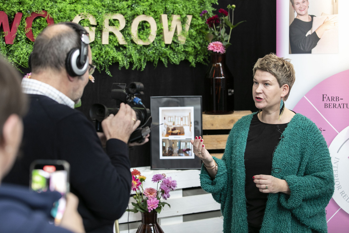 Eine Frau hält einen Vortrag vor ihrem Messestand und wird dabei von einem Kameramann und einer Frau mit dem Handy gefilmt.