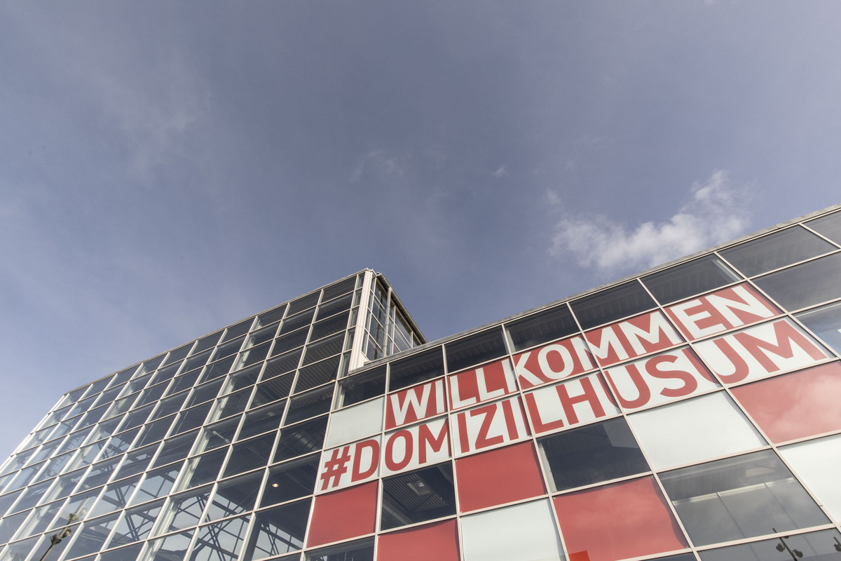 Der Bildausschnitt zeigt ein von unten fotografiertes Messegebäude mit dem Banner Willkommen #Domizilhusum in den Fenstern.