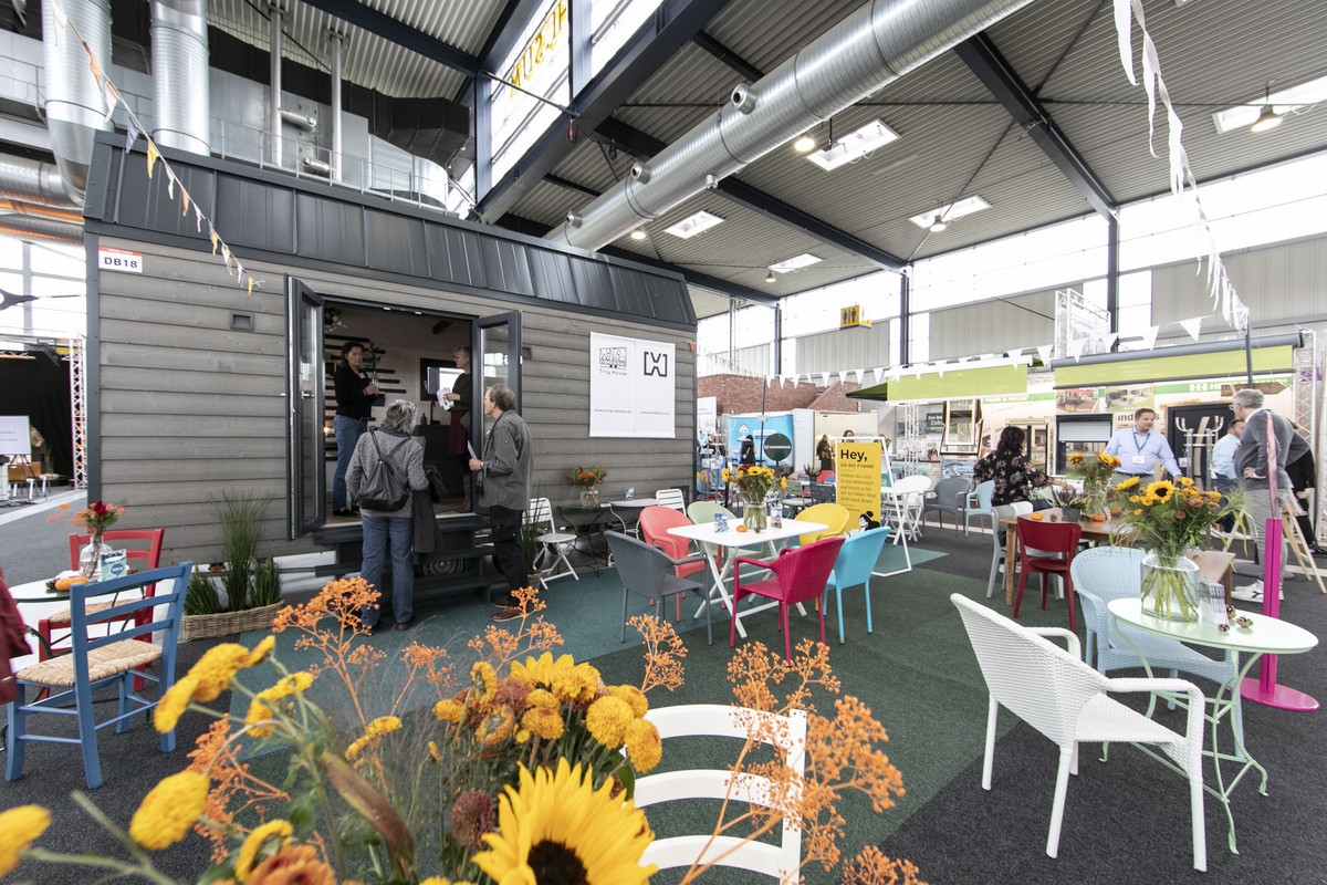 Ein Messestand mit Tischen und Stühlen davor. Auf den Tischen stehen Blumenvasen mit Sonnenblumen darin.