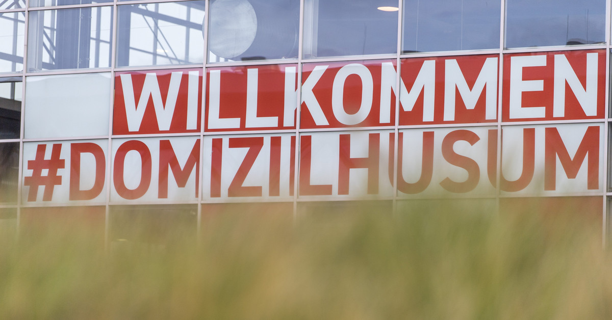 Ein Bildausschnitt, der einen kleinen Teil der Messefenster mit der Banneraufschrift Willkommen #Domizilhusum zeigt. Davor befinden sich unscharfe Grashalme.